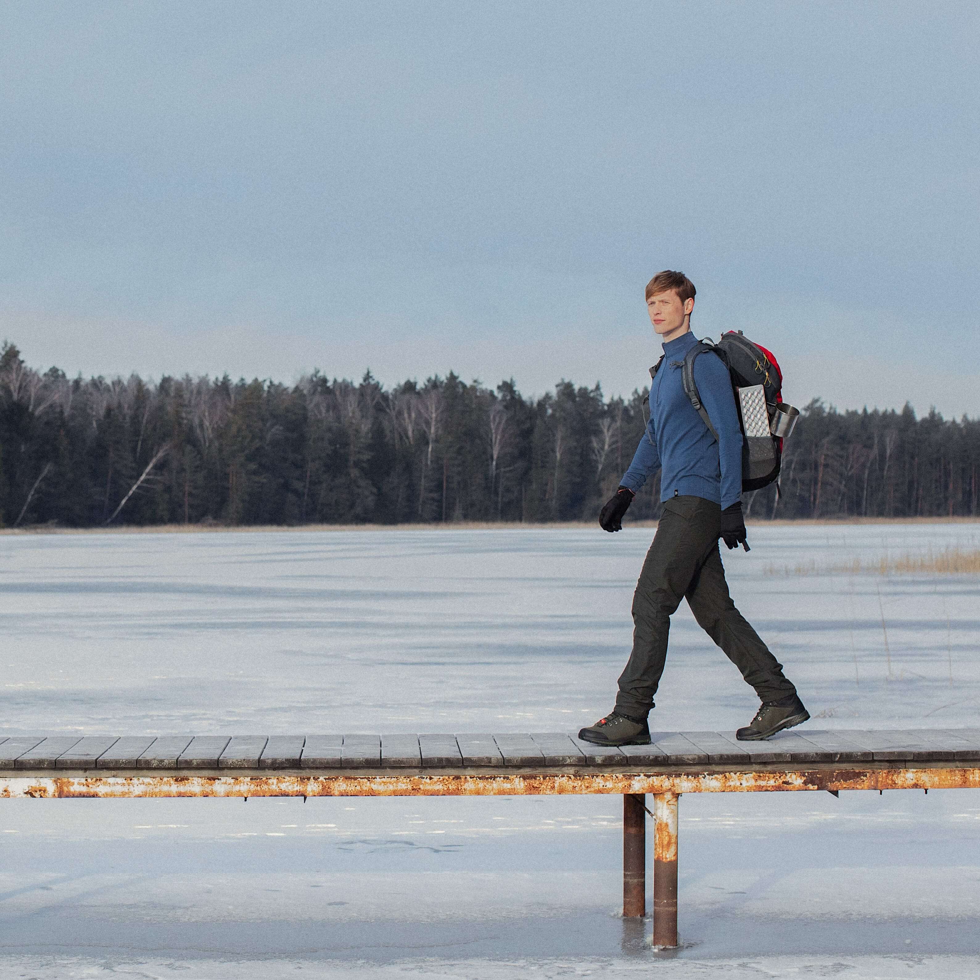 a-man-with-a-backpack-walks-on-a-bridge-over-a-frozen-lake-in-a-forest